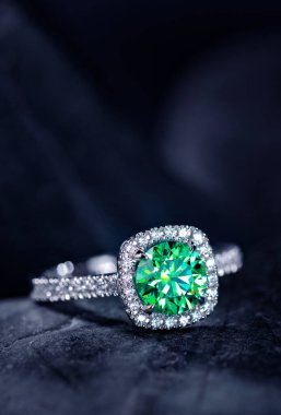 Beyaz bantlı yeşil elmas bir yüzük siyah bir yüzeye oturur. Yüzük, lüks ve zarif bir görünüm kazandıran elmaslardan bir halkayla çevrilidir. Yeşil arasındaki kontrastı.