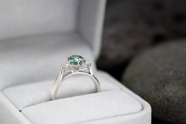 Güzel yeşil ve beyaz bir yüzük beyaz bir kutunun içinde. Yüzük altından yapılmış ve ortasında bir elmas var. Yüzük görüntünün ana odak noktasıdır.