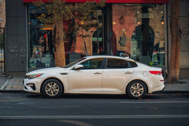 Beyaz bir KIA arabası yolun kenarına park edilmiş bir binanın önünde. Araba bir pencerenin önüne park edilmiş ve binanın önünde duran bir kişi var.