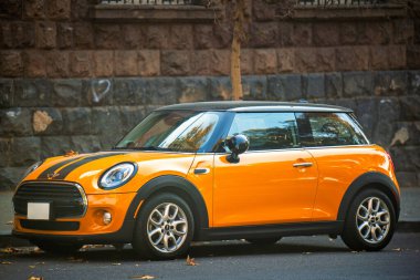 Parlak turuncu bir mini Cooper caddenin kenarına park etmiş. Araba tuğla bir duvarın önünde park edilmiş ve yerde yapraklar var. Araba, park etmesi kolay küçük, kompakt bir araçtır.