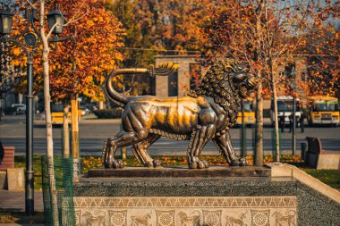 Bir binanın önünde bir aslan heykeli duruyor. Heykel bronzdan yapılmıştır ve altın yelesi vardır. Heykel ağaçlar ve kaldırımlarla çevrili.