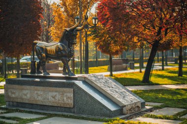 Kafasında lamba olan bir boğa heykeli parktadır. Heykel ağaçlar ve banklarla çevrili.