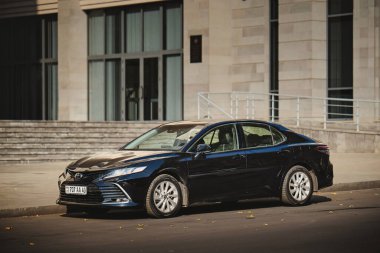 Siyah bir araba caddenin kenarına park edilmiş bir binanın önünde. Araba bir Toyota Corolla.