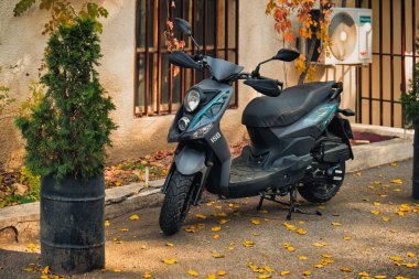 Siyah bir scooter bir binanın önündeki kaldırımda park halinde. Yerdeki yapraklar sarı ve kahverengidir. Sahneye sonbahar havası katar.