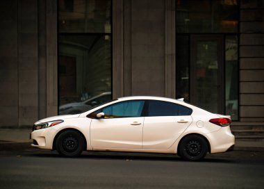 Beyaz bir araba bir binanın önündeki bir caddeye park edilmiştir. Araba küçük bir sedan ve bir park yerinde park halinde.