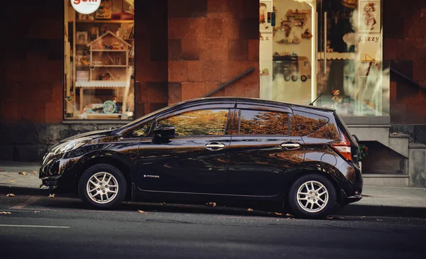 Siyah bir araba bir binanın önündeki caddede park halinde. Araba yolun kenarına park edilmiş ve kaldırımda duran bir kişi var. Sahne sessiz ve sakin, arabayla.