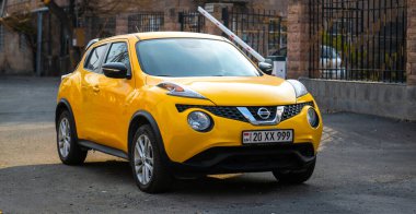 MX-99 plakalı sarı bir Nissan arabası bir otoparkta duruyor. Araba bir çitin önünde park edilmiş.