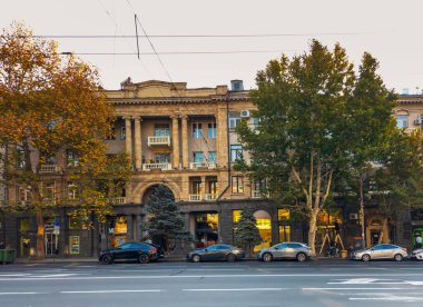 Erivan Ermenistan 11.25.2025. Arka planda büyük bir bina olan bir sokak manzarası. Sokak ağaçlarla dolu ve kaldırım boyunca park edilmiş birkaç araba var.