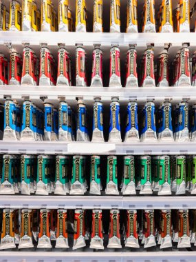 Rafa dizilmiş bir sıra boya tüpü var. Tüpler farklı renk ve boyutlarda.