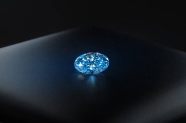 Siyah bir yüzeyde mavi bir elmas duruyor. Elmas parıldıyor, odadaki ışığı yansıtıyor.