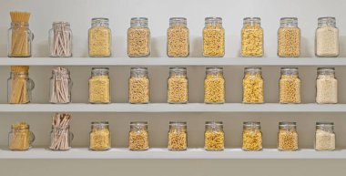 Farklı renkte makarnadan yapılmış cam kavanozlarla dolu bir raf. Kavanozlar, içinde spagetti ve makarna bulunan kavanozlarla sıralanmış. Bolluk ve çeşitlilik kavramı