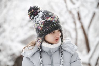 Şapka ve ceket giyen bir kız çocuğu karda duruyor. Kafasında kabarık bir şapka ve boynunda bir eşarp var.