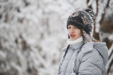 Şapka ve ceket giyen bir kız çocuğu karda duruyor. O bir eşarp ve beyaz bir şapka giyiyor.