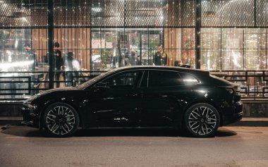 Siyah bir araba bir binanın önündeki caddede park halinde. Araba bir pencerenin önünde park edilmiş ve binanın dışında bekleyen insanlar var. Sahne karanlık.