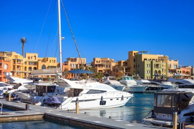 El Gouna Mısır. Birçok teknesi olan bir yat limanı. Tekneler, mavi çizgili beyaz bir tekne de dahil olmak üzere çeşitli boyutlarda ve renklerde. Gökyüzü açık ve mavi
