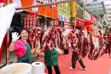 Chongqing Çin 'de bir kadın bir et standının önünde yüzünde bir gülümsemeyle duruyor. Tavandan sarkan birkaç parça et var ve kadın önlük giyiyor. Sahne canlı görünüyor.