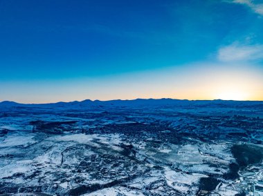 Birkaç bulutlu mavi bir gökyüzü ve arka planda karla kaplı bir dağ. Gökyüzü açık ve güneş batıyor.