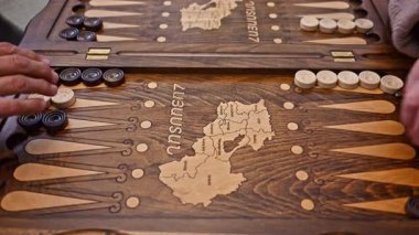 Yaşlı bir adam ve yaşlı bir kadının elleri tavla oynuyor, adam zar atıyor ve altılı alıyor, sonra da dama oynatıyorlar.