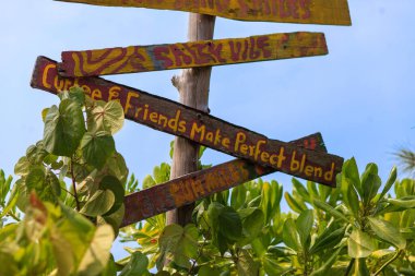 Canlı işaretler yıpranmış bir tahta direğe dayanır. Elle boyanmış kelimeler tropikal cazibe ve samimi titreşimleri yansıtır. Yemyeşil yapraklar kırsal işaret direğini çerçeveliyor. Güneşli gökyüzü sıcak, davetkar bir zemin sunar