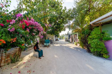 Çiçeklerle kaplı huzurlu köy şeridi. Kadın sessizce oturur, sakin düşünceler içinde kaybolur. Yumuşak güneş ışığı yukarıdaki yapraklı dalların arasından süzülür. Tozlu yol, gezginleri sessiz köşeleri keşfetmeye davet eder.