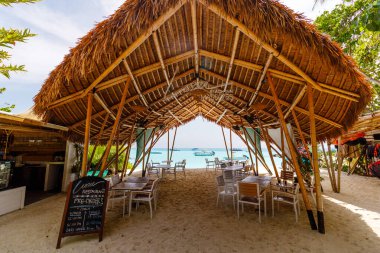 Kumlu kumsal, sazdan yapılma çatı sığınağıyla tanışır. Tahta kirişler sıcak ve doğal bir örtüyü destekler. Sessiz yemek ve rahatlama için masa sandalyeleri ayarlandı. Yumuşak güneş yeşil yapraklar ve açık gökyüzünde parlar.