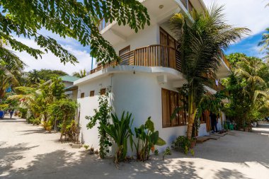 Ahşap balkonları ve tropik yeşillikleri olan beyaz bir bina. Güneş ışığı yaprakların arasından kumlu zemine süzülür. Sıcak, davetkar ve huzurlu ada inzivası havası. Seyahat ve tatil için mükemmel.