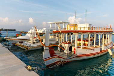 Geleneksel tekne sakin liman sularında dinlenir. Parlak gökyüzü nazik, turkuaz dalgaları yansıtır. Tekne, açık güverteler ve tahta korkuluklardan oluşuyor. Sakin bir atmosfer, sakin bir yolculuk ya da rahatlık...