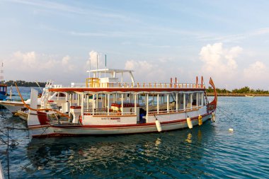 Beyaz tekne turkuaz sularda yüzüyor. Yumuşak bulutlar bulutların üzerinde süzülür. Sıcak ışık, yumuşak deniz kıyısında yıkanır. Yolcular üst güvertede dinleniyor. Tekneler kıyı şeridinde demirli.