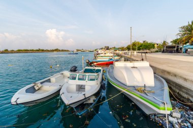 Sakin sular, sessiz bir rıhtıma demirlemiş tekneleri yansıtır. Beyaz ve yeşil gemiler açık mavi bir gökyüzünün altında dinleniyor. Uzak adalar sakin liman sahnesini çerçeveliyor. Durgun yüzeyde güneş ışığı parlar