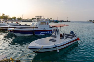 Tekne sakin liman sularında sakince dinlenir. Yumuşak güneş ışığı, yumuşak mavi denizin üzerinde parlar. Kırmızı çizgili beyaz gövde ince bir kontrast ekler. Daha büyük bir geminin yanında, huzurlu bir manzara.