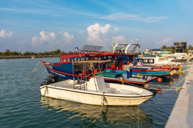 Renkli tekneler sakin liman sularında huzur içinde dinleniyor. Güneş ışığı sakin deniz yüzeyinde yavaşça parlar. Balıkçı tekneleri, küçük ve büyük, rıhtım tarafında. Gökyüzü dağınık bir şekilde genişliyor