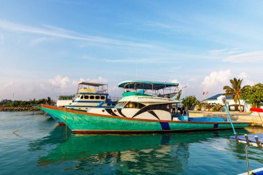 Turkuaz tekne kıyıya yakın sularda yüzüyor. Mavi gölgelikler üst güvertede konfor için. Parlak gökyüzü ve uzak palmiye ağaçları sahnesi. Canlı renkler güneşli deniz yüzeyini yansıtır