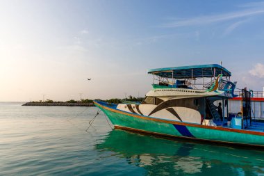 Renkli tekne sakin turkuaz sularda yüzer. Mavi gölgelik güvertedeki yolcuları gölgeliyor. Uzak ada ufkunda hafif bir ışık parlıyor. Sakin deniz gökyüzünü ve teknenin parlak renklerini yansıtır.