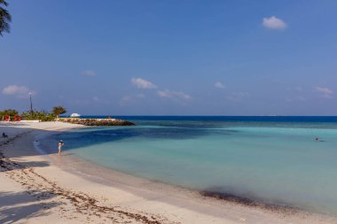 Yumuşak beyaz kum ve turkuaz suları olan güneşli bir sahil. Yalnız figür, sakin ve rahat bir şekilde kıyı şeridinde geziniyor. Uzak ufukta ince bulutlu açık mavi gökyüzü