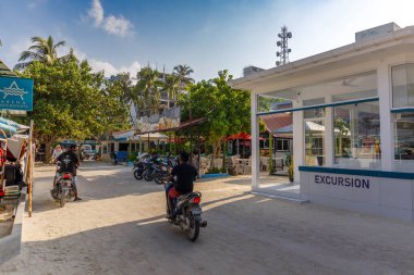 Motosikletler tropik bir köy caddesinde süzülüyor. Palmiye ağaçlarının gölgesinde, sahne sıcak ve canlı görünüyor. 