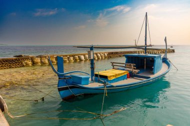 Mavi balıkçı teknesi taştan dalgalanma suyuyla sakin duruyor. Güneş ışığı turkuaz suların üzerinde altın gibi parlar. Kıyı sahnesi sükunet ve sonsuz cazibe yayar. Seyahat ve denizcilik için ideal.