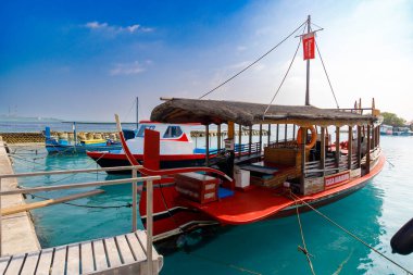 Kırmızı ahşap tekne turkuaz sularda yüzer. Çatı, yolcuları güneşten ve meltemden korur. Kablo hatları yukarı doğru uzanıyor ve kırsal cazibe ekleniyor. Güneşli gökyüzü parlak, sakin denizi yansıtıyor.
