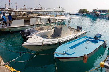 Temiz turkuaz su beşikleri demirli tekneleri sakinleştirir. Beyaz ve mavi gemiler hareketli bir limanda huzur içinde dinleniyor. İki adam rıhtımda durup suyu izliyor. Güneş ışığı yüzeyden yansıyor.