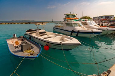 Güneşli tekneler sakin turkuaz sularda dinlenir. İpli halatlar, taş iskelenin yakınındaki demir damarları. Açık gökyüzü ve uzak toprak huzur derinliği ekler. Teknelerin boyutları küçük ve orta büyüklüktedir.
