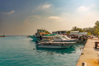 Turkuaz sular sakin bir limanı kucaklıyor. Tekneler yumuşak, puslu gökyüzünün altında dinlenir. Canlı yeşil palmiyeler sahne çerçevesi. İnsanlar iskelenin yanında toplanır, rahat ve kaygısızdır.