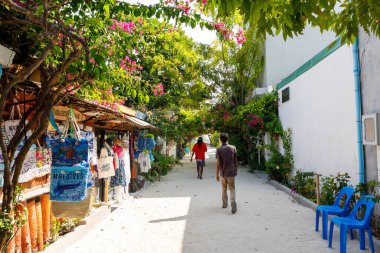 Bereketli ağaçlar, güneşli, sessiz bir ara sokağı çerçeveler. Tepemizde parlak pembe çiçekler açar ve renk katar. İki insan kendi düşüncelerinde kaybolarak dolaşır. El yapımı mallar soldaki kırsal bir tezgahta asılı.