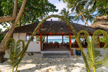 Sazlı çatısı ve ahşap masaları olan tropik bir sahil kulübesi. Kalp şeklinde palmiye yaprakları girişi çerçeveliyor. Kırmızı fenerler açık hava sığınağının altında asılı. Mavi okyanus dalgaları yavaşça arkadan çarpar