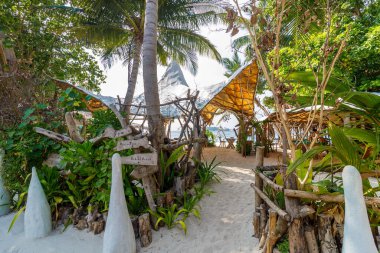 Tropikal yol yemyeşil sularda esiyor. Beyaz heykeller kumlu bir geçidi çerçeveliyor. Ahşap barınaklar palmiyelerin altında gölge sunar. Doğa kırsal, sakin tasarım ile harmanlanır. Tatil için mükemmel bir yer.