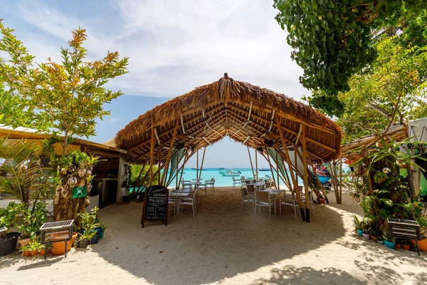 Sazlı çatı altında tropik bir sahil restoranı. Yumuşak güneş ışığı yeşil yaprakların arasından süzülür. Beyaz kum, turkuaz deniz ufkuna kadar uzanıyor. Sandalyeler ve masalar sessiz yemek için hazır.