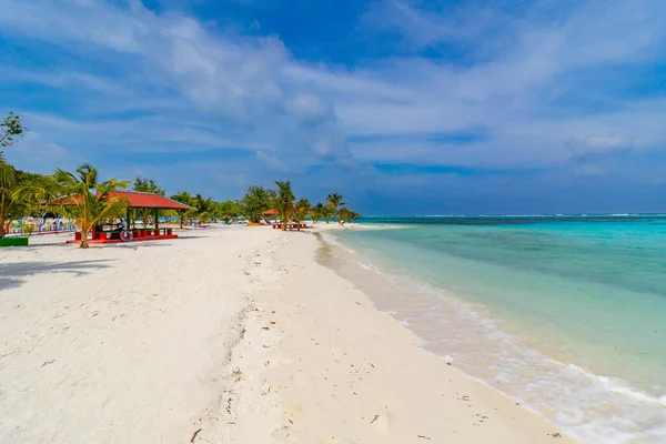 Yumuşak kum, parlak mavi gökyüzünün altında turkuaz dalgalarla buluşur. Palmiye ağaçları, kırmızı çatılı bir sahil sığınağının yanında usulca sallanıyor. Sakin plaj rahatlamaya ve sessizce kaçmaya davet eder.