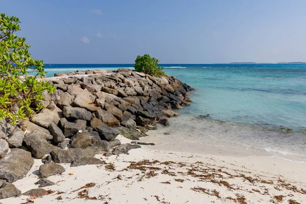 Güneş ışığı, kayalık kıyıların yanındaki turkuaz sularda parıldıyor. Yeşillik, engebeli ve doğal bir kıyı şeridini çerçeveler. Dalgakıran yakınında beyaz kum yosunlarıyla harmanlanır.