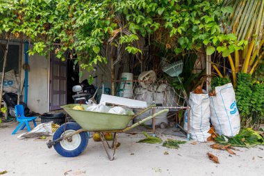 Yeşil yapraklı sarmaşıklar kırsal bir el arabasını gölgeliyor. Mavi el arabası dağınık bir kapının yanında duruyor. Kum ve yapraklar zemini dağıtır. Plastik sandalye yakınlarda duruyor.