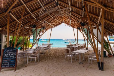 Sazdan çatı sığınağı olan tropik bir sahil kafesi. Beyaz sandalyeler örülmüş tavanın altındaki kumlu kıyılarda. Açık mavi okyanus, açık uzayın ötesine uzanır. Güneş ışığı doğal olarak süzgeçten geçer.