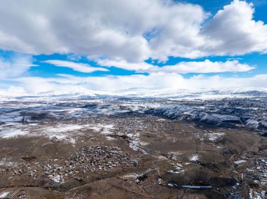 Kar, açık mavi gökyüzünün altında, tüylü beyaz bulutlarla kaplı uzak tepeleri örtüyor. Engebeli arazide ve dağınık binalarda güneş ışığı parlıyor. Huzurlu kış manzarası sükuneti davet eder