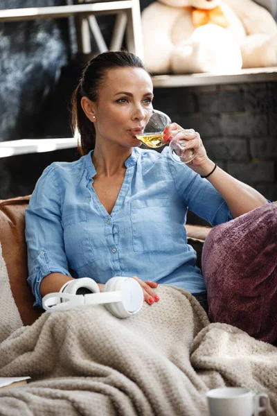 Έννοια ελεύθερου χρόνου. Ευτυχισμένη όμορφη γυναίκα πίνει λευκό κρασί από γυαλί κάθεται σε έναν καναπέ σε εσωτερικούς χώρους. Γυναίκα ξοδεύει την ελεύθερη ημέρα της και χαλαρώνει στο σπίτι μόνη της. — Φωτογραφία Αρχείου